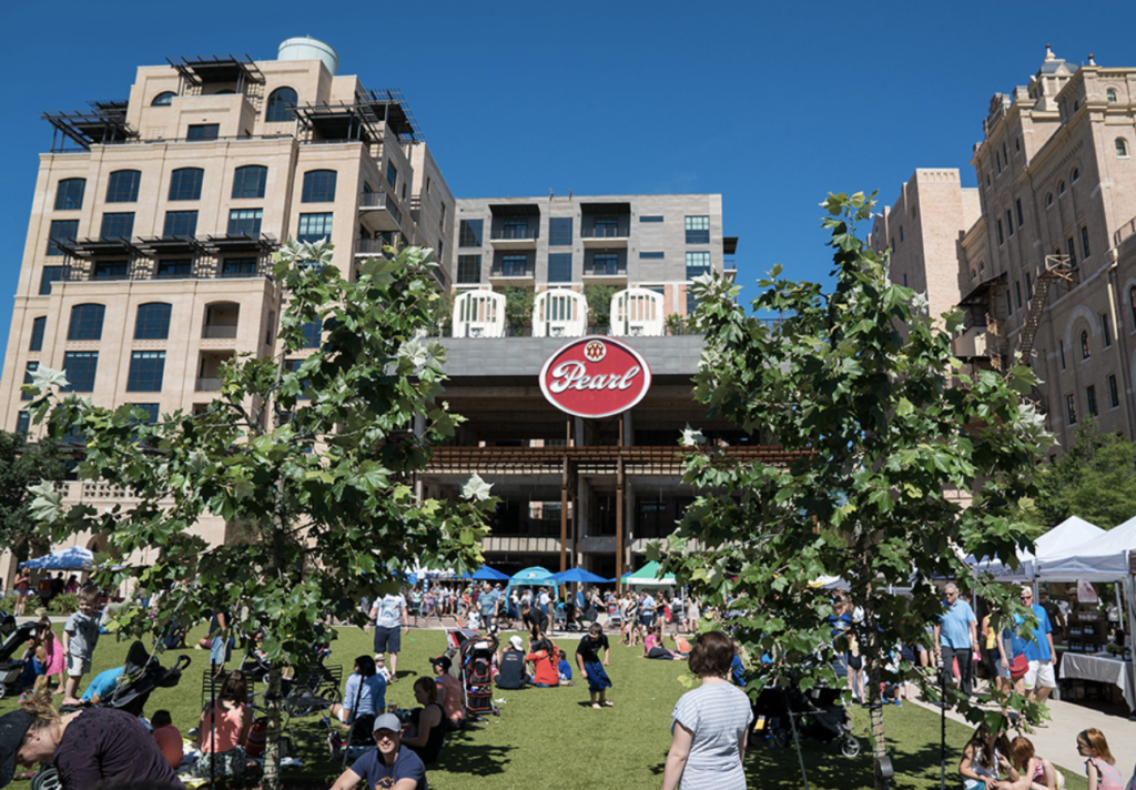 People congregate in a field of synthetic grass in front of a building with a sign that reads "Pearl." Tall buildings are in the background.